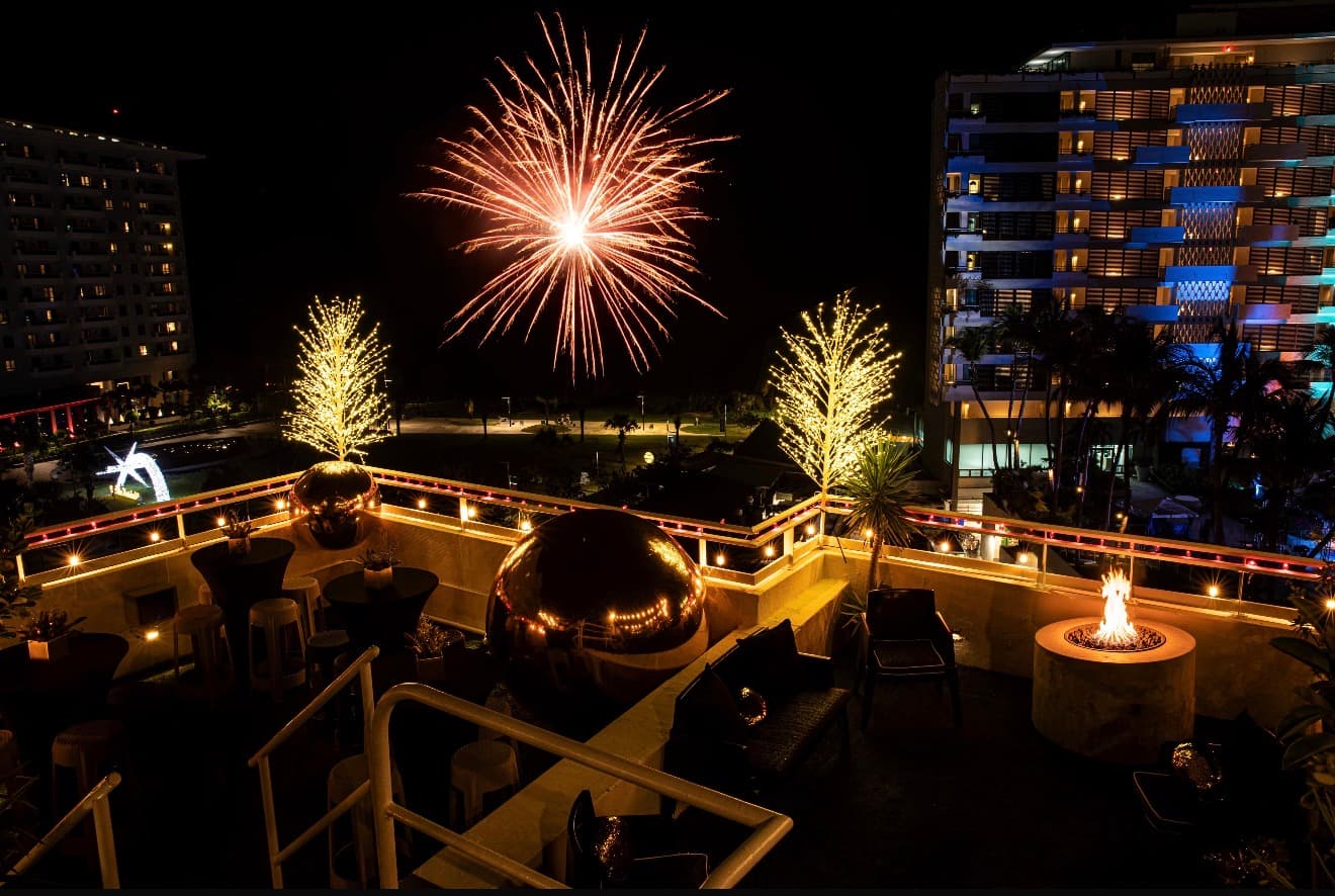 Fireworks over a rooftop celebration with golden string lights and a fire pit